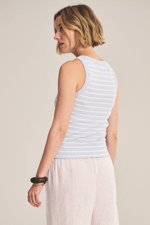 A woman with short blonde hair models the MORIE TANK TOP by Velvet by Graham & Spencer—a light blue and white striped cotton rib fitted sleeveless top—paired with white pants, standing slightly turned against a plain background.