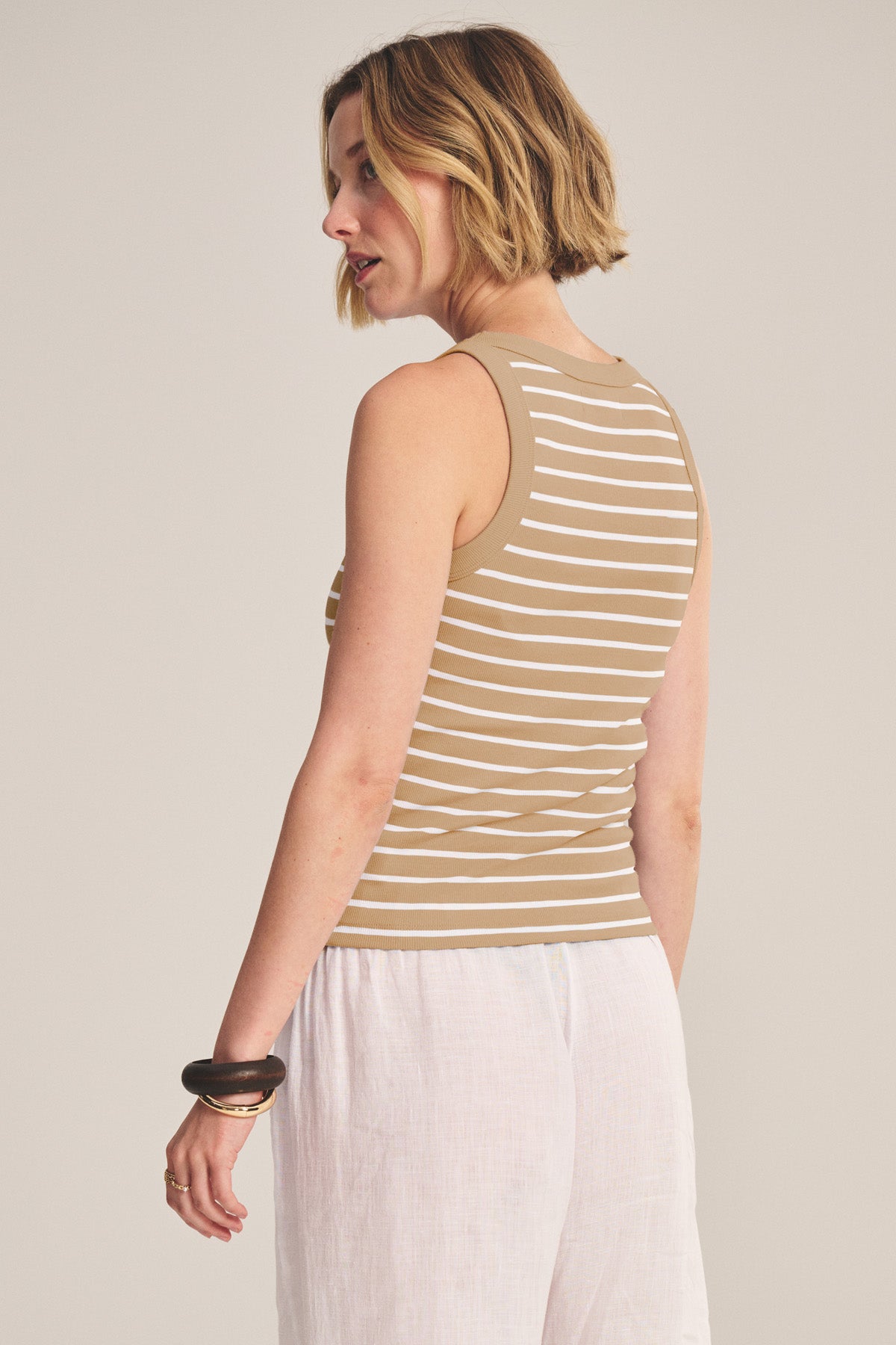 A woman in the MORIE TANK TOP by Velvet by Graham & Spencer—a beige and white striped cotton cropped tank with a fitted silhouette—pairs it with white pants and stands facing slightly away from the camera.