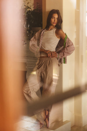 A woman stands indoors, one hand on her hip and looking to the side, wearing the Velvet by Graham & Spencer BROOKS PANT with a parachute silhouette, paired with a striped tank top and patterned open shirt.