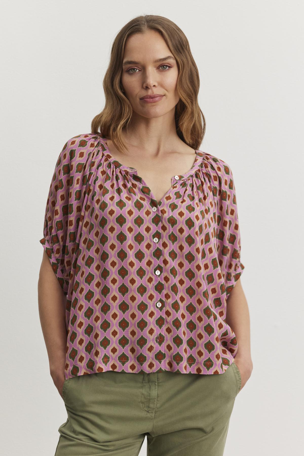 A woman with wavy hair wears the HARA TOP by Velvet by Graham & Spencer alongside green pants, standing with hands in pockets against a plain background.-38483587104961