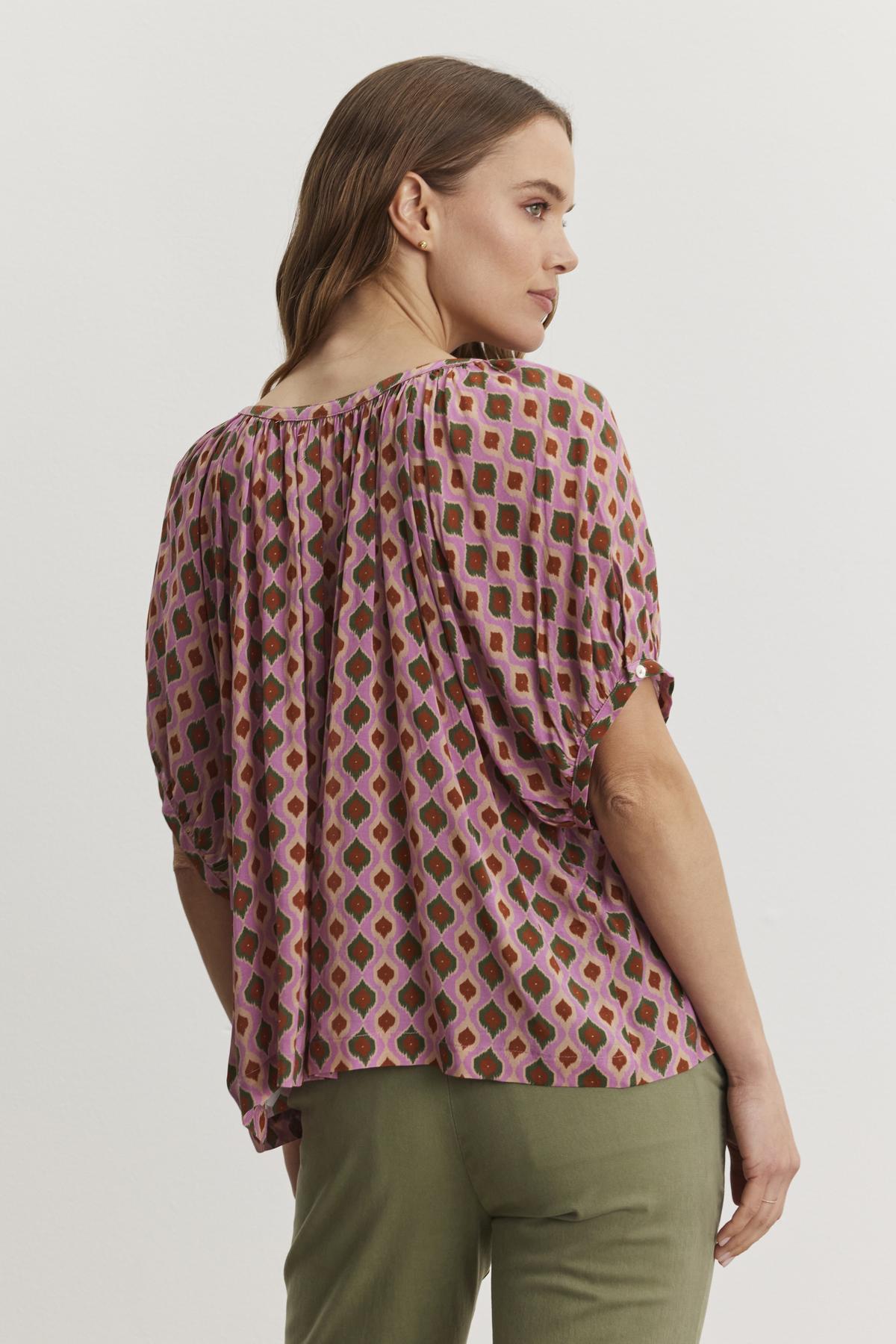   A woman with long hair is wearing the HARA TOP from Velvet by Graham & Spencer, which features a Moroccan print with pink and green shapes, paired with green pants. She is facing away, looking to the side. 