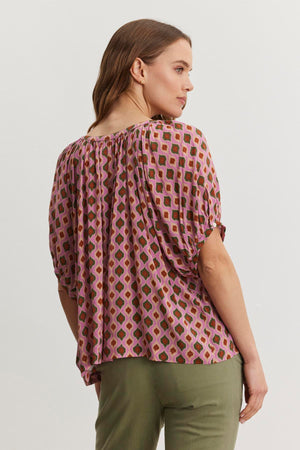 A woman with long hair is wearing the HARA TOP from Velvet by Graham & Spencer, which features a Moroccan print with pink and green shapes, paired with green pants. She is facing away, looking to the side.