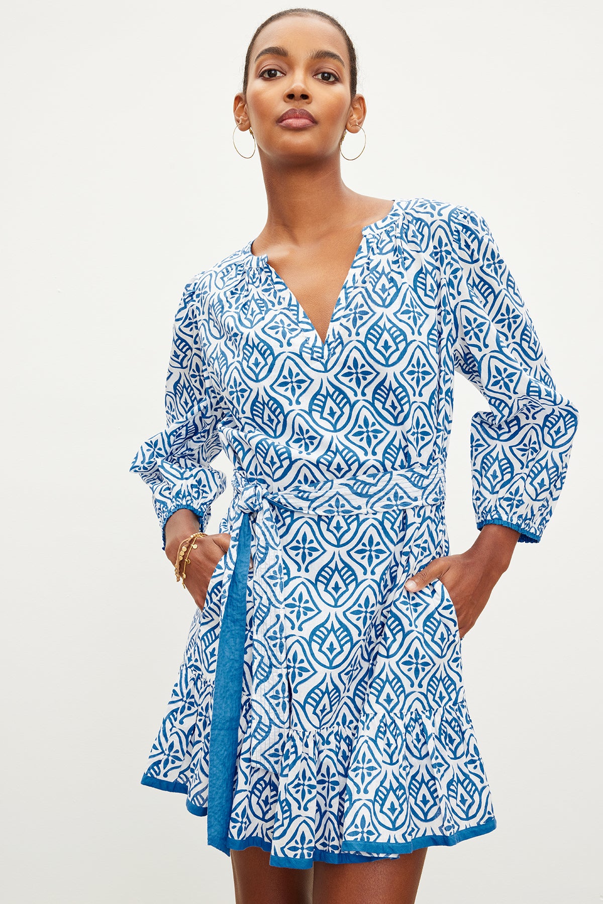 A person stands wearing the Velvet by Graham & Spencer KENLEY DRESS, a blue and white mosaic-printed cotton dress with a v-neckline and long sleeves, hands in pockets.-35967628935361