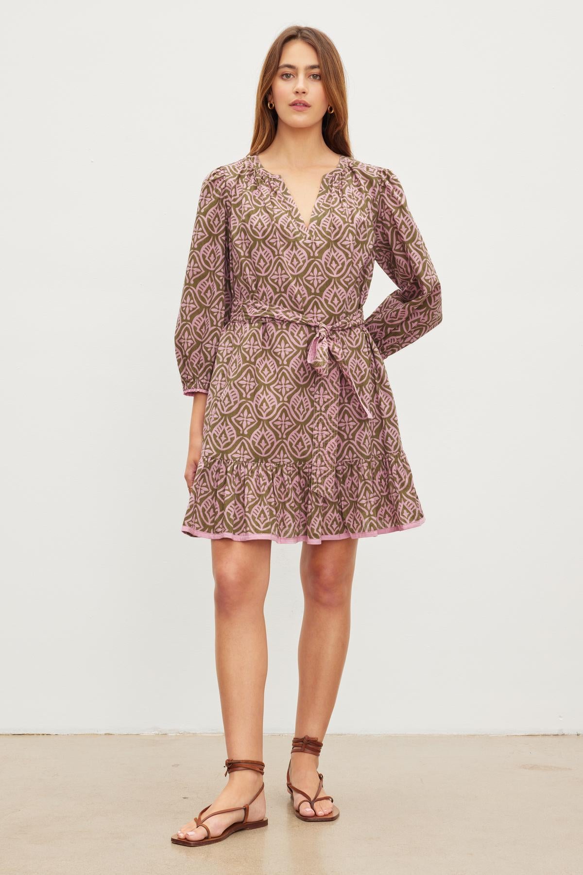 A woman wearing a Velvet by Graham & Spencer KENLEY DRESS, a mosaic-printed, long-sleeve, V-neckline dress with a sash belt, stands against a plain white background. She is also wearing brown sandals.-37073710186689