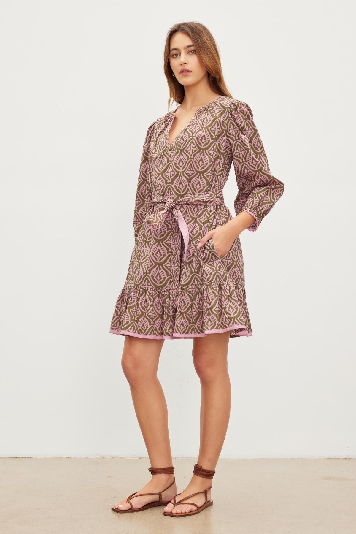   A woman stands in a Velvet by Graham & Spencer KENLEY DRESS, a mosaic-printed cotton, belted A-line dress with long sleeves and a v-neckline, wearing flat sandals. She has her hands in the dress pockets and stands against a plain white background. 