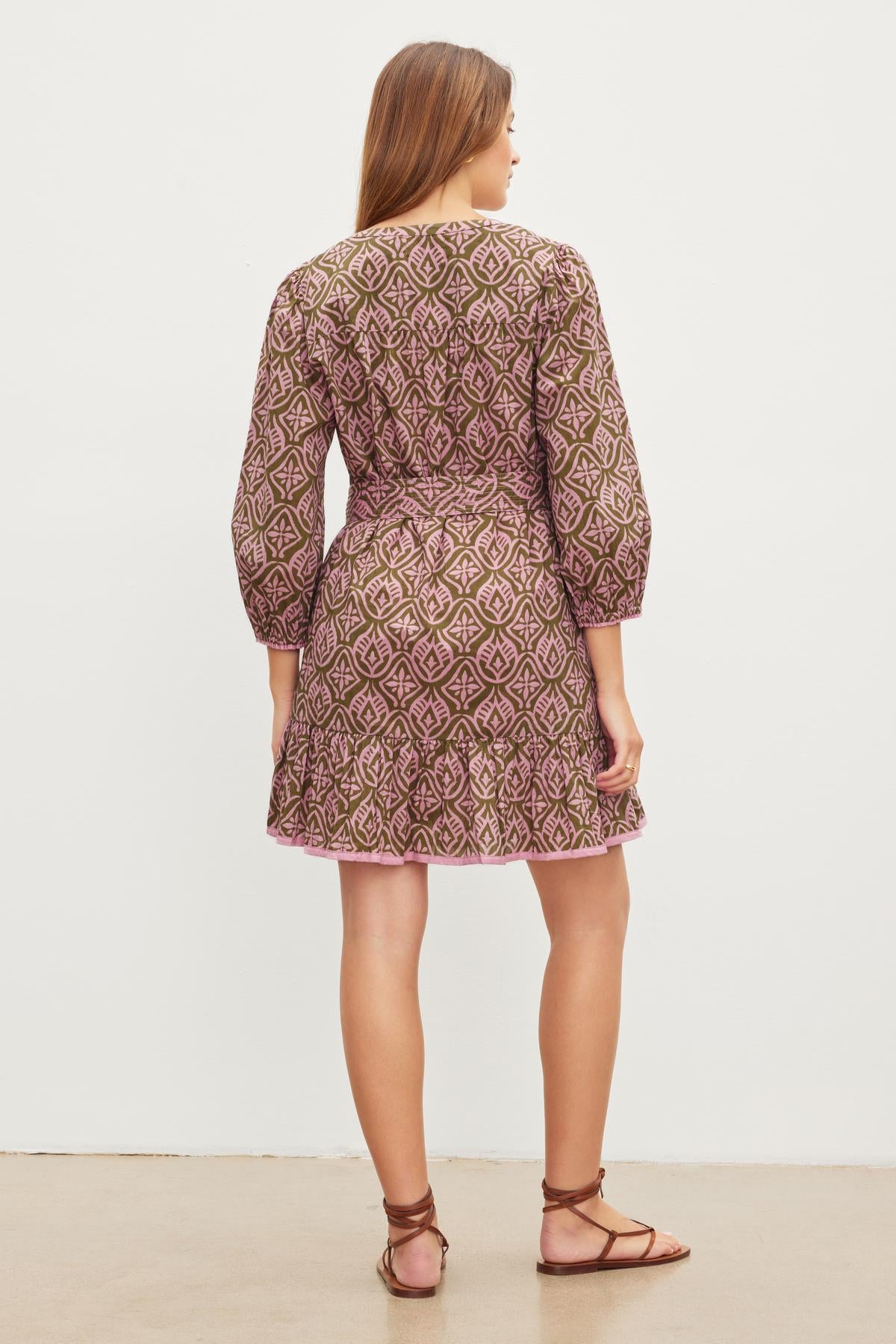 A woman with long hair stands facing away, wearing the Velvet by Graham & Spencer KENLEY DRESS, a mosaic-printed cotton A-line dress with three-quarter sleeves and a v-neckline, paired with sandals, against a plain white background.-37073710284993