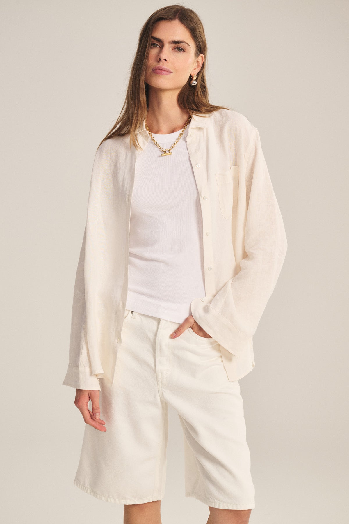 A woman wears the MULHOLLAND LINEN SHIRT by Velvet by Jenny Graham—styled loosely over a white tank and knee-length shorts—standing against a plain light background.-41044038746305