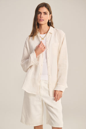 A woman wears the MULHOLLAND LINEN SHIRT by Velvet by Jenny Graham, styled with a white top and knee-length shorts, standing against a plain background.