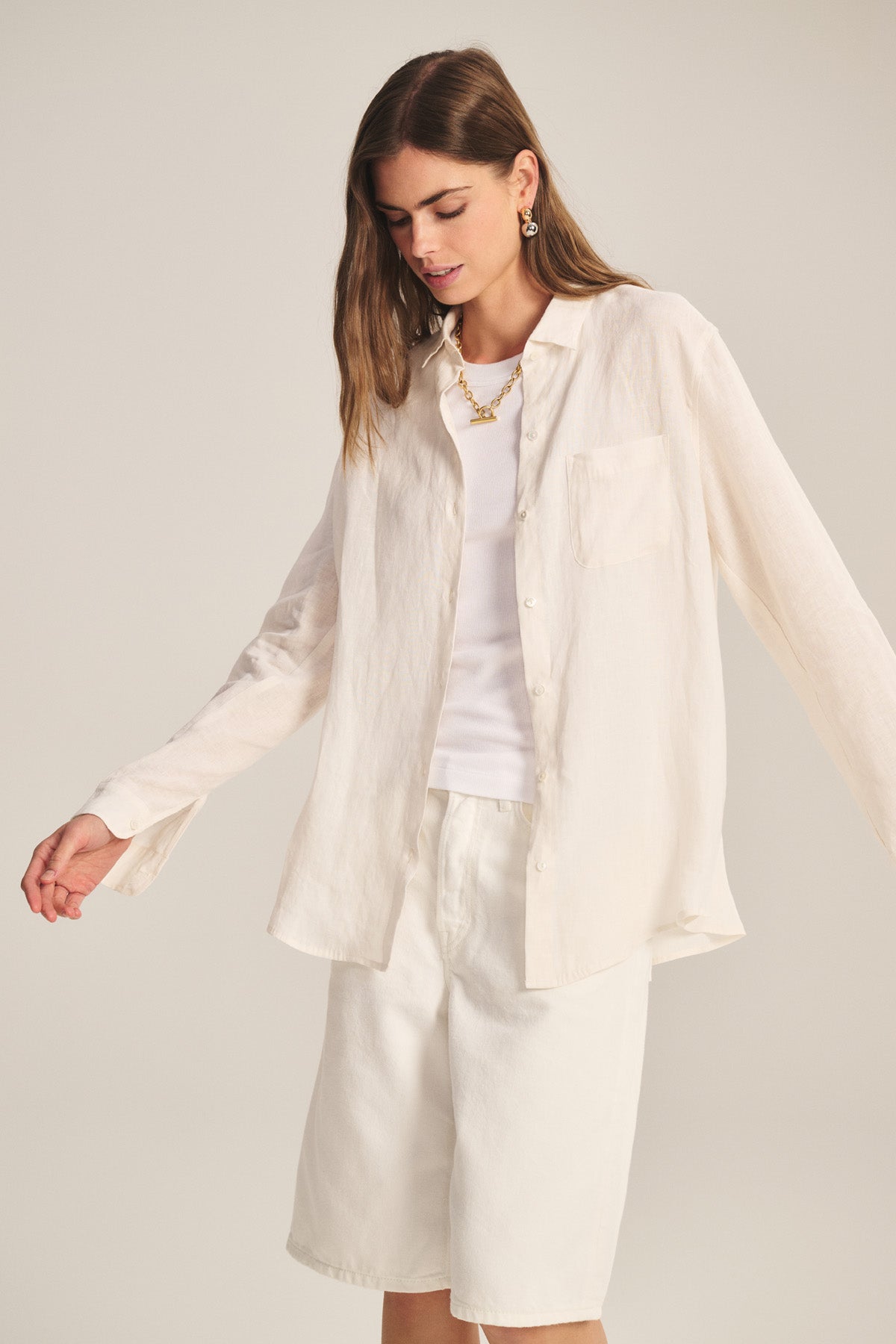 A woman in the MULHOLLAND LINEN SHIRT by Velvet by Jenny Graham, layered over a white tank and paired with knee-length white shorts, stands against a neutral background.-41044038680769