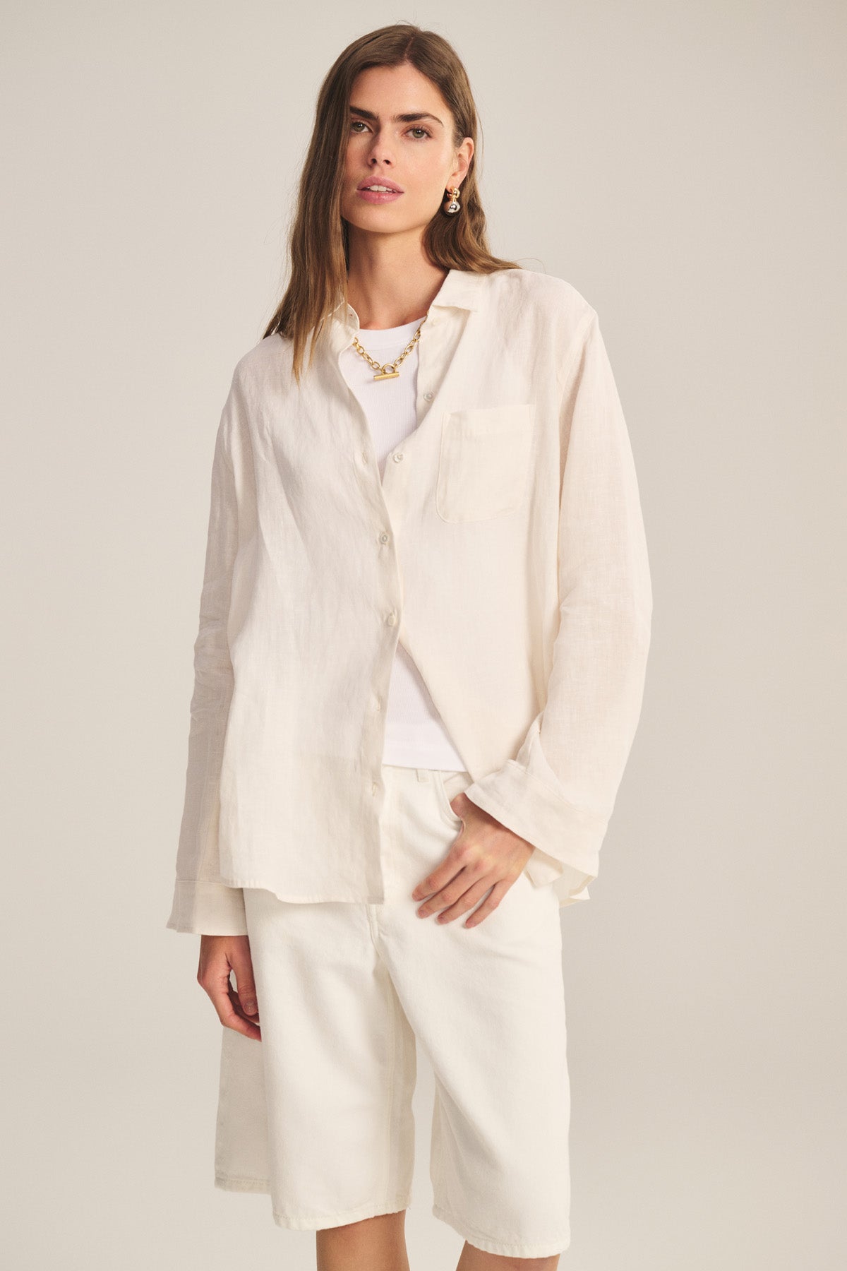 A woman wears the MULHOLLAND LINEN SHIRT by Velvet by Jenny Graham, a loose light beige button-up with a relaxed silhouette, layered over a white top and knee-length white shorts against a plain background.-41044038648001