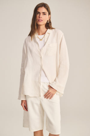 A woman wears the MULHOLLAND LINEN SHIRT by Velvet by Jenny Graham, a loose light beige button-up with a relaxed silhouette, layered over a white top and knee-length white shorts against a plain background.