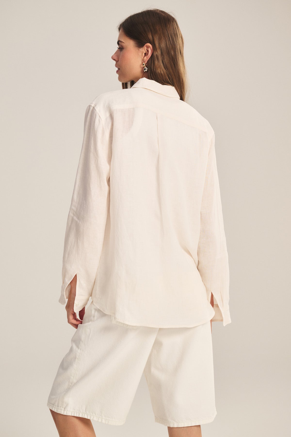 A woman stands with her back to the camera, wearing the Velvet by Jenny Graham MULHOLLAND LINEN SHIRT featuring a relaxed silhouette and scooped hemline, paired with knee-length cream shorts against a plain light background.