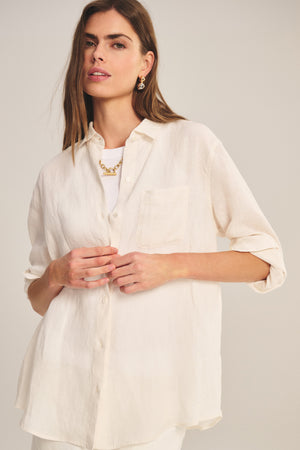 A woman poses against a neutral background wearing the MULHOLLAND LINEN SHIRT by Velvet by Jenny Graham—a loose-fitting, cream linen button-up with rolled sleeves and a relaxed silhouette—layered over a white top.