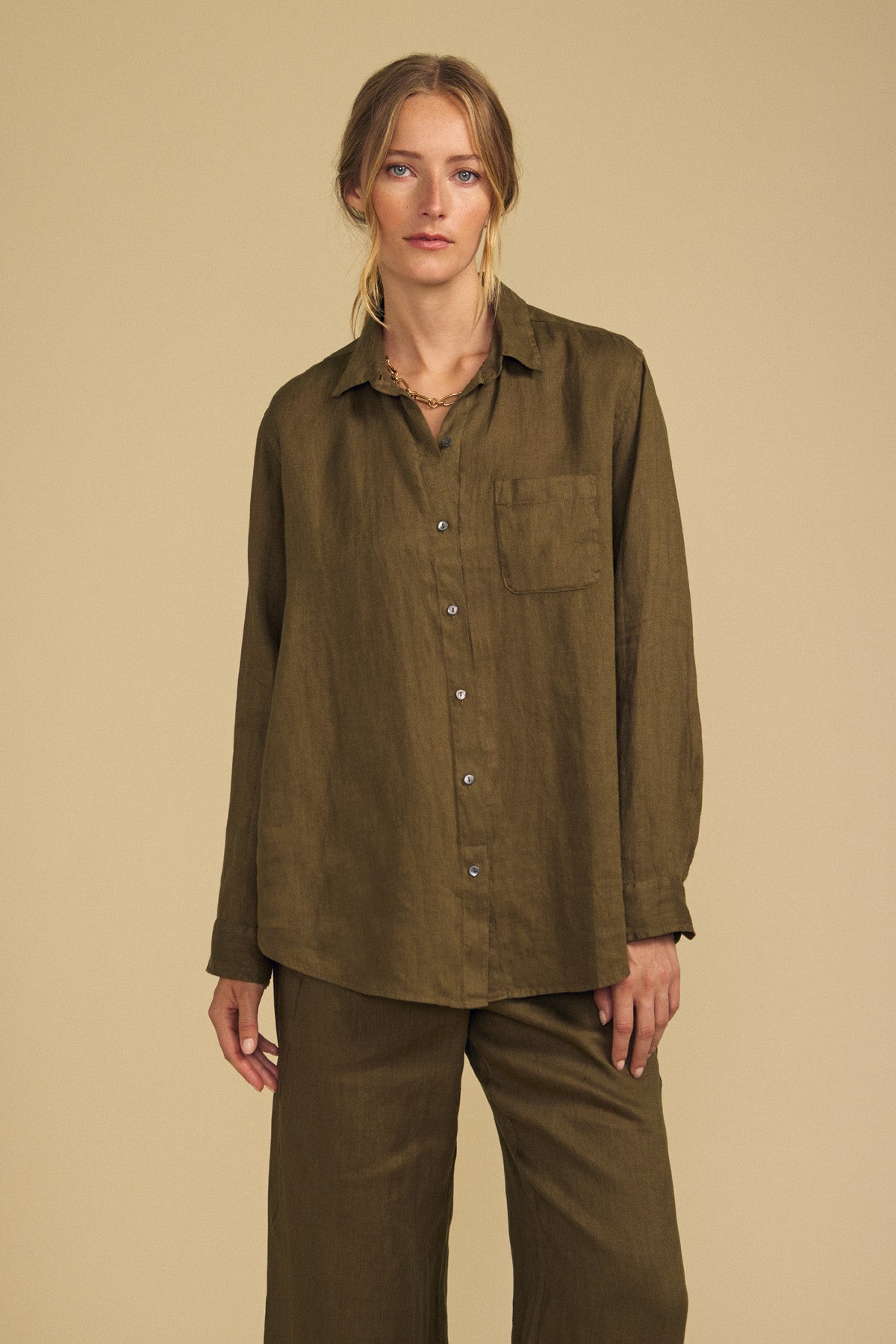 A person wearing the MULHOLLAND LINEN SHIRT by Velvet by Jenny Graham, featuring a relaxed silhouette in olive green with a chest pocket and matching pants, stands against a beige background.
