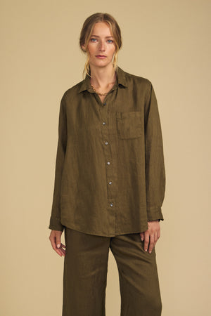 A person wearing the MULHOLLAND LINEN SHIRT by Velvet by Jenny Graham, featuring a relaxed silhouette in olive green with a chest pocket and matching pants, stands against a beige background.