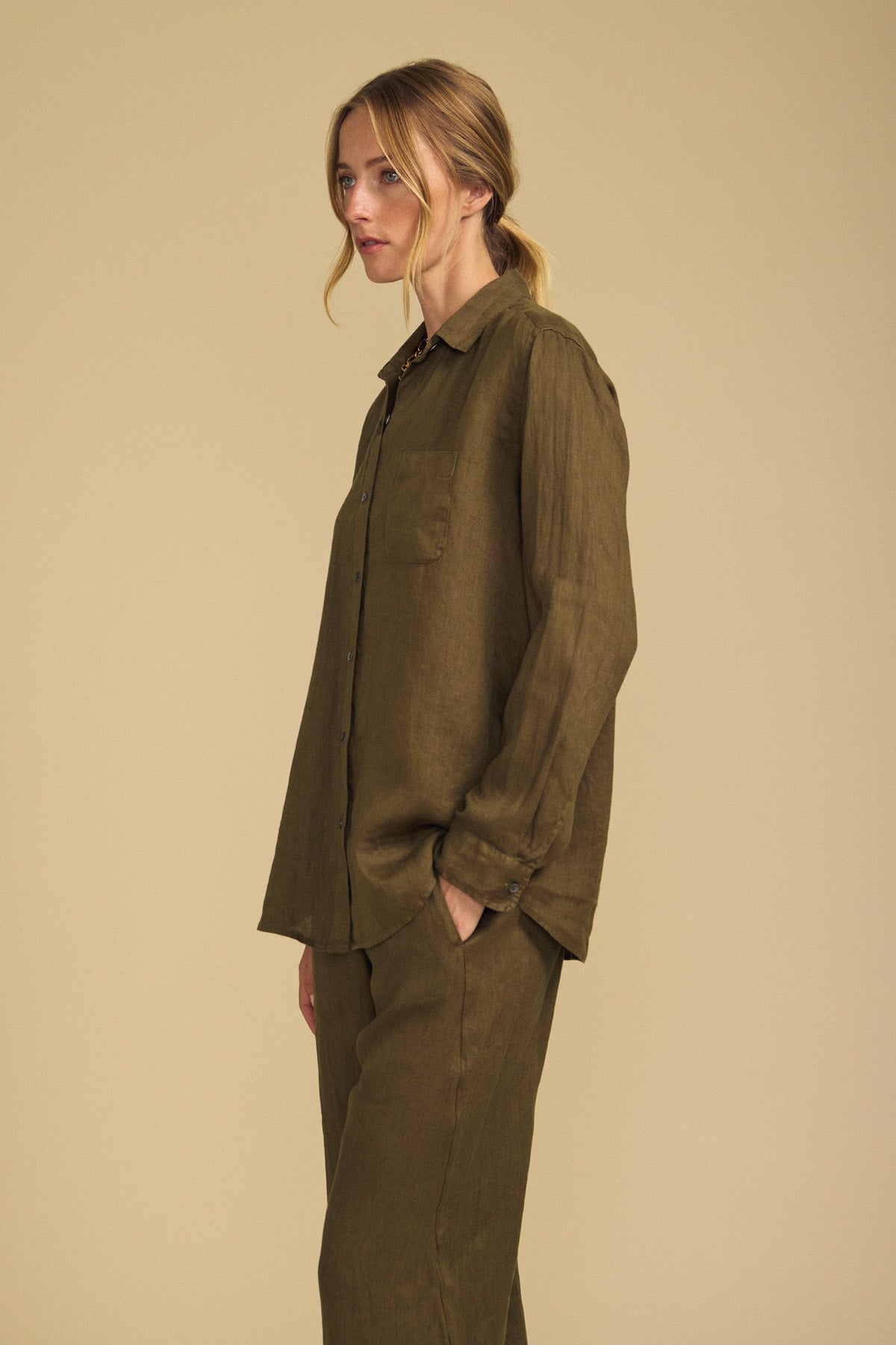 A woman models the MULHOLLAND LINEN SHIRT by Velvet by Jenny Graham in olive green, paired with matching pants. Standing against a beige background with her hands in her pockets, she showcases the shirt’s relaxed silhouette that brings effortless style and comfort to her look.-41838212645057