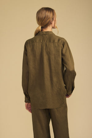 A person with blonde hair in a low ponytail stands against a beige background, facing away from the camera, wearing the MULHOLLAND LINEN SHIRT and matching pants by Velvet by Jenny Graham in olive green. The relaxed silhouette of this breathable linen ensemble exudes effortless style and comfort.