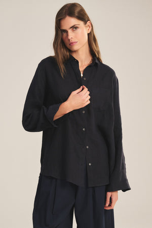 A woman with long brown hair wears the Velvet by Jenny Graham MULHOLLAND LINEN SHIRT, a loose-fitting button-up with a scooped hemline, and matching black pants, standing against a neutral background.