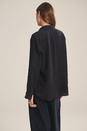 A person with long brown hair, wearing the MULHOLLAND LINEN SHIRT by Velvet by Jenny Graham and black pants, stands facing away from the camera.