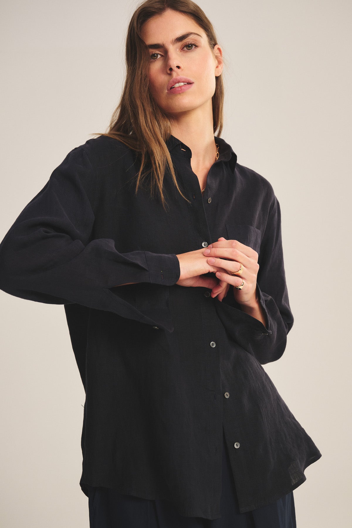 A person with long brown hair wears the Velvet by Jenny Graham MULHOLLAND LINEN SHIRT, a loose-fitting black linen button-up with a scooped hemline, standing against a neutral background and looking at the camera with a neutral expression.