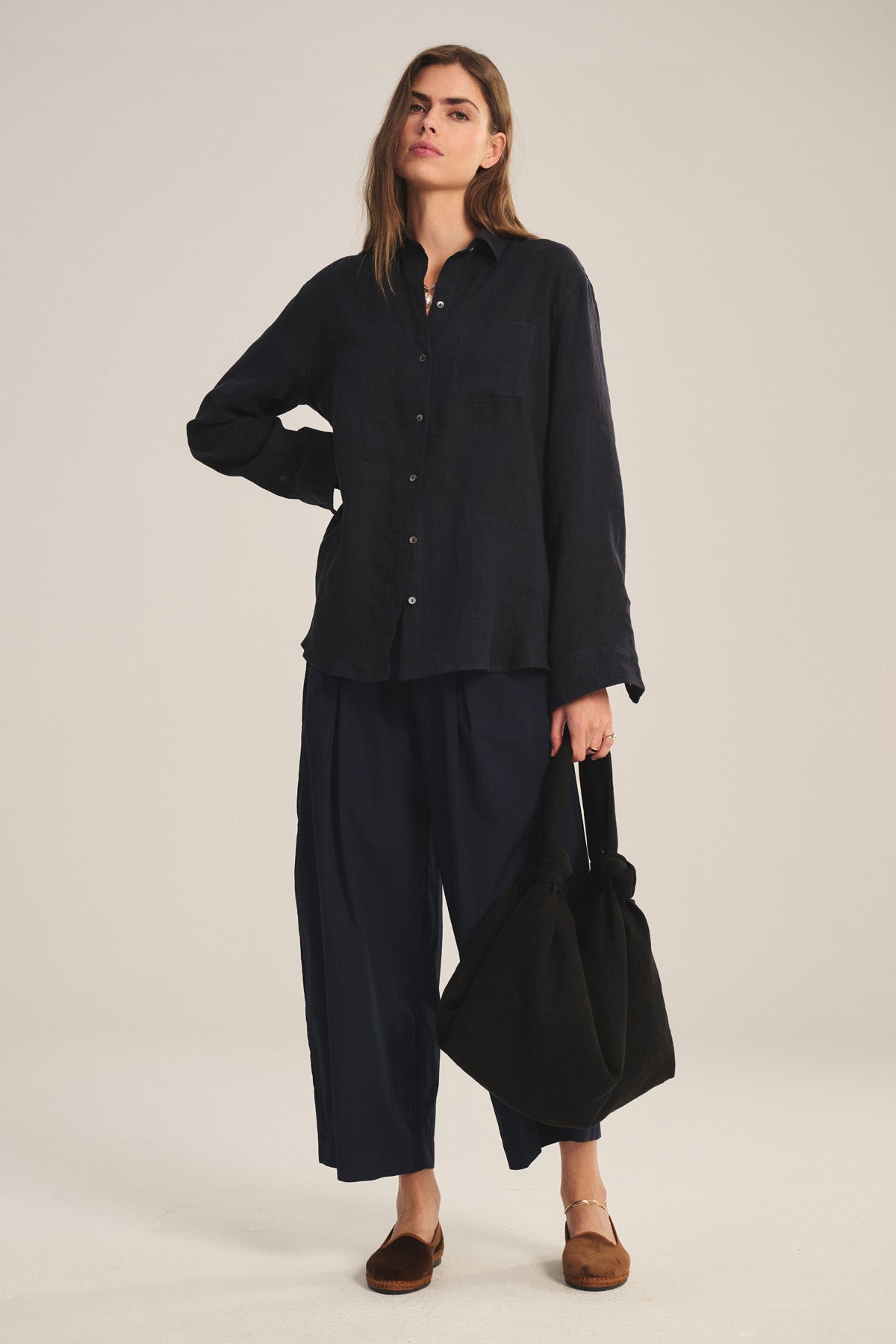 A woman stands with one hand on her hip, wearing a loose dark shirt, Velvet by Graham & Spencer’s EASTON PANT—modern wide-leg dark pants with an elastic waist—brown flats, and holding a large black bag.-41037963362497