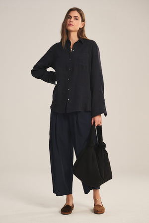 A woman stands with one hand on her hip, wearing a loose dark shirt, Velvet by Graham & Spencer’s EASTON PANT—modern wide-leg dark pants with an elastic waist—brown flats, and holding a large black bag.