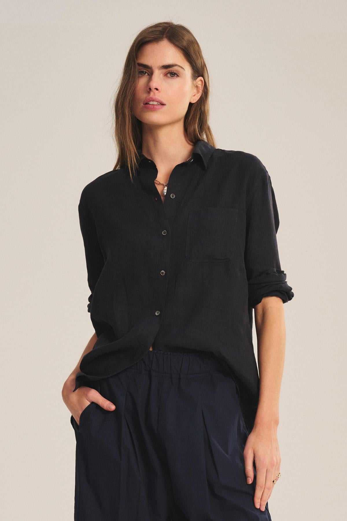A person stands against a neutral background, wearing the MULHOLLAND LINEN SHIRT by Velvet by Jenny Graham with sleeves rolled up and dark pants. The shirt’s relaxed silhouette complements their casual pose, with one hand in their pocket.