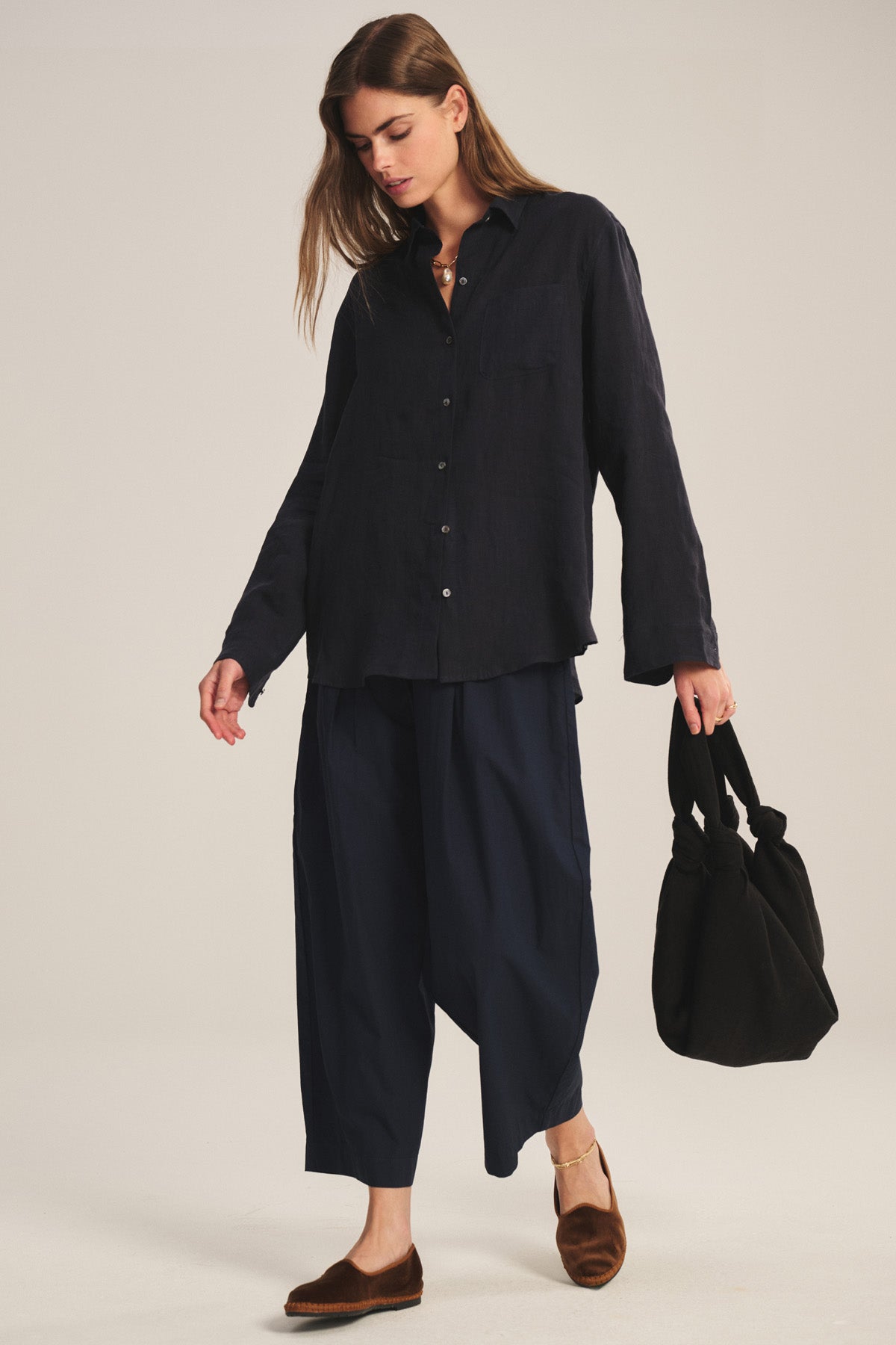 A woman stands against a neutral background, wearing the EASTON PANT by Velvet by Graham & Spencer—wide navy elastic waist pants—paired with a loose dark button-up shirt and holding a black bag while looking down.