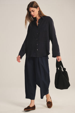 A woman stands against a neutral background, wearing the EASTON PANT by Velvet by Graham & Spencer—wide navy elastic waist pants—paired with a loose dark button-up shirt and holding a black bag while looking down.