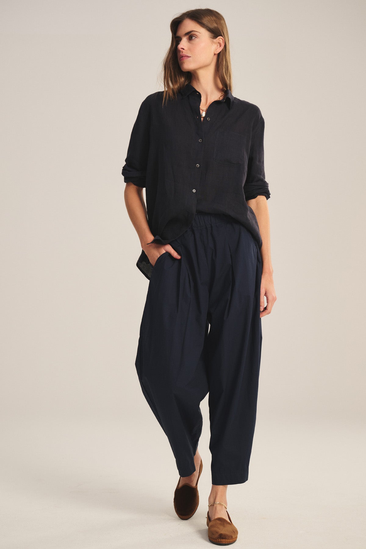 A person stands against a plain background wearing the EASTON PANT by Velvet by Graham & Spencer—cropped pants with an elastic waist—paired with a dark button-up shirt and brown flats, one hand in their pocket.