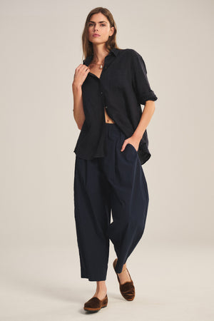 A woman stands against a plain background wearing the EASTON PANT by Velvet by Graham & Spencer—an elastic waist, wide navy cropped pant paired with a loose dark button-up shirt and brown flats, embodying true modern wardrobe essentials.