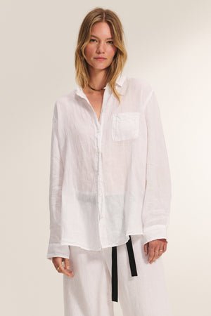 A woman stands against a plain light background, wearing the MULHOLLAND LINEN SHIRT by Velvet by Jenny Graham—a relaxed silhouette, long-sleeved linen button-up—paired with white pants.