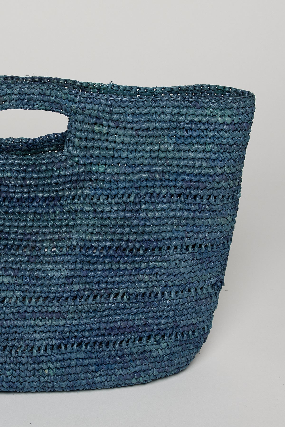   A close-up view of a textured, blue NAOMI HANDHELD STRAW BAG from Velvet by Graham & Spencer with a prominent handle. 