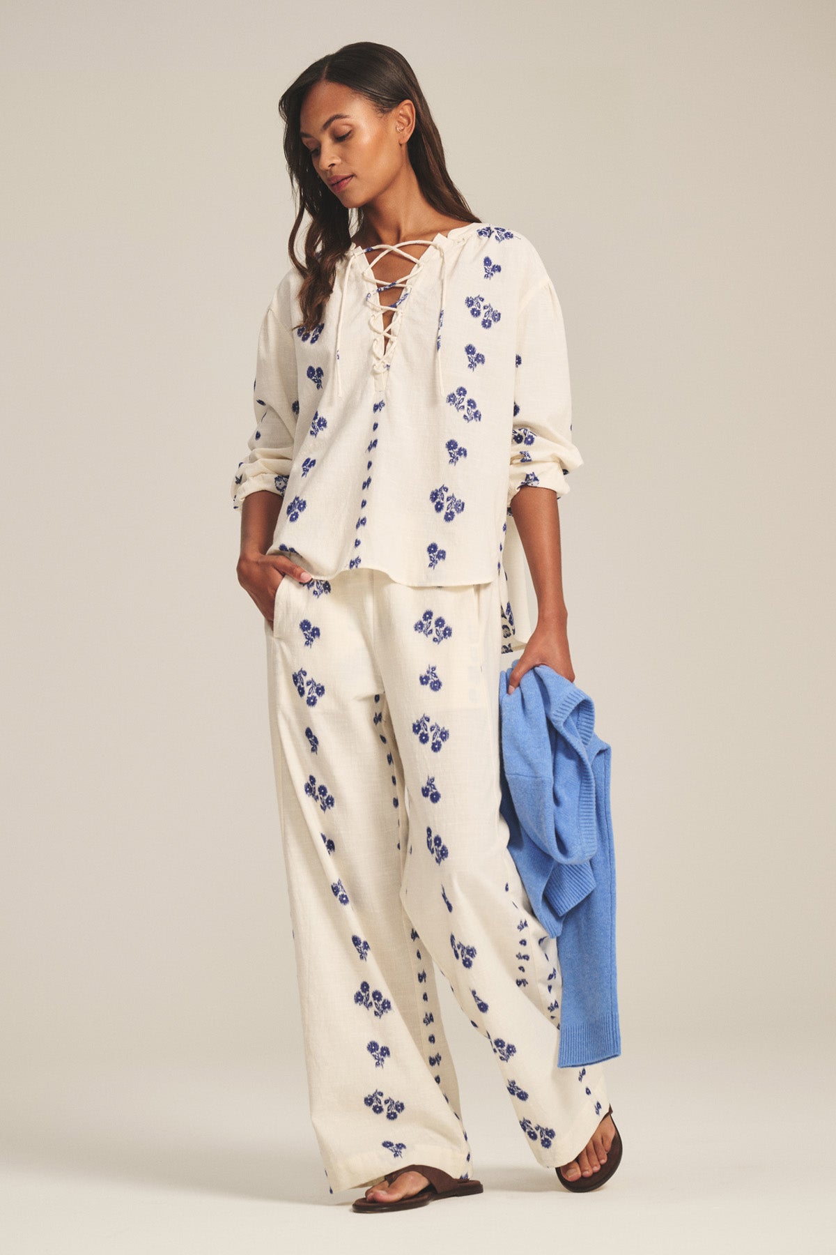 A woman stands against a plain background wearing the NELLIE PANT by Velvet by Graham & Spencer—wide-leg cotton pants with jacquard stitching and an elastic waist—paired with an ivory laced blouse and holding a blue garment.-41338483343553
