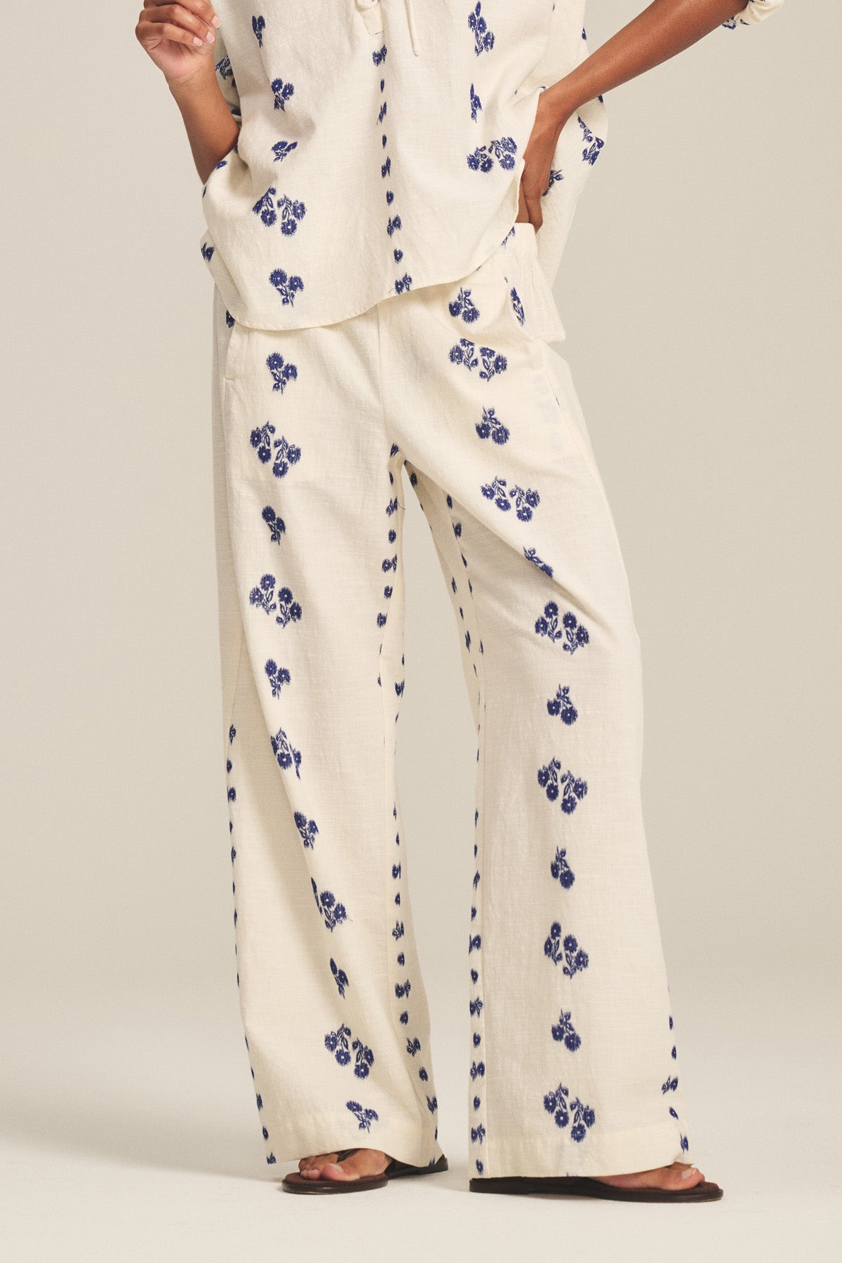 A person wears the NELLIE PANT by Velvet by Graham & Spencer—cream wide-leg jacquard cotton pants with blue floral prints and an elastic waist—paired with a matching top and brown sandals, standing against a plain background.-41338483376321