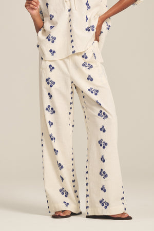 A person wears the NELLIE PANT by Velvet by Graham & Spencer—cream wide-leg jacquard cotton pants with blue floral prints and an elastic waist—paired with a matching top and brown sandals, standing against a plain background.