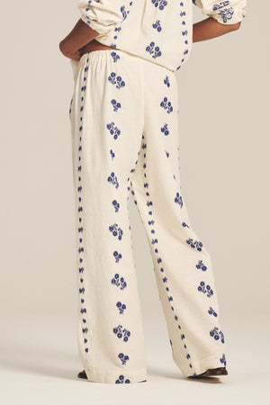 Model wearing the NELLIE PANT by Velvet by Graham & Spencer—cream jacquard cotton wide-leg pants with blue floral print and elastic waist. Matching top partially visible; standing with hand on hip against a plain background.