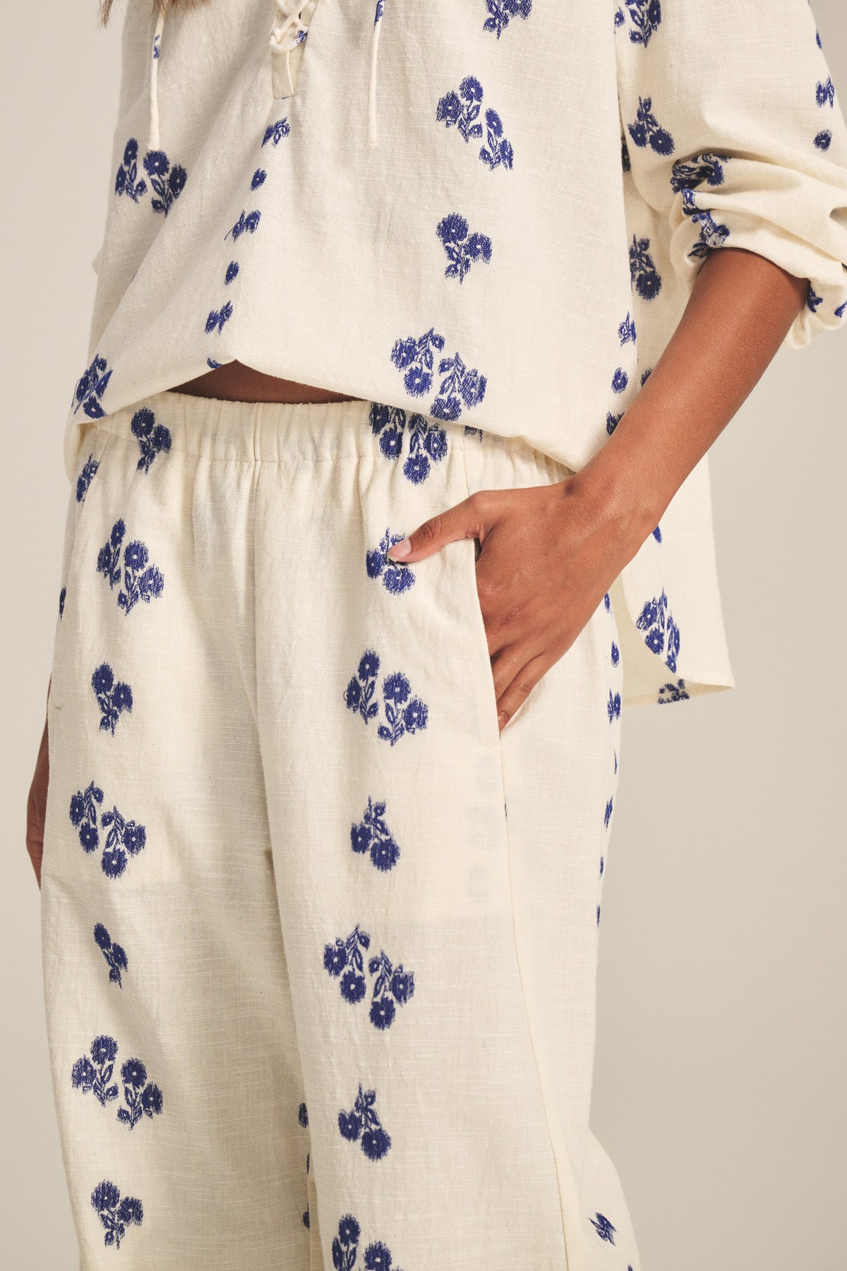 A person models the NELLIE PANT by Velvet by Graham & Spencer—a cream jacquard cotton set with blue floral prints, featuring a long-sleeve top and elastic waist wide-leg pants, with one hand in the pocket.-41338483441857