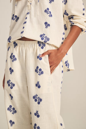 A person models the NELLIE PANT by Velvet by Graham & Spencer—a cream jacquard cotton set with blue floral prints, featuring a long-sleeve top and elastic waist wide-leg pants, with one hand in the pocket.