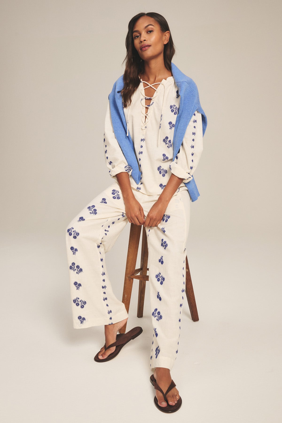 Woman wears the NELLIE PANT by Velvet by Graham & Spencer—wide-leg cotton pants with a pull-on elastic waist—paired with a cream floral top, black flip-flops, and a blue sweater draped over her shoulders while sitting on a stool.-41338483146945