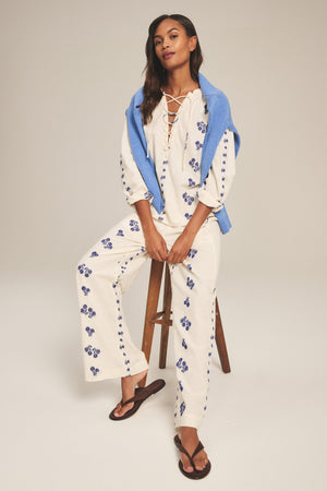 Woman wears the NELLIE PANT by Velvet by Graham & Spencer—wide-leg cotton pants with a pull-on elastic waist—paired with a cream floral top, black flip-flops, and a blue sweater draped over her shoulders while sitting on a stool.