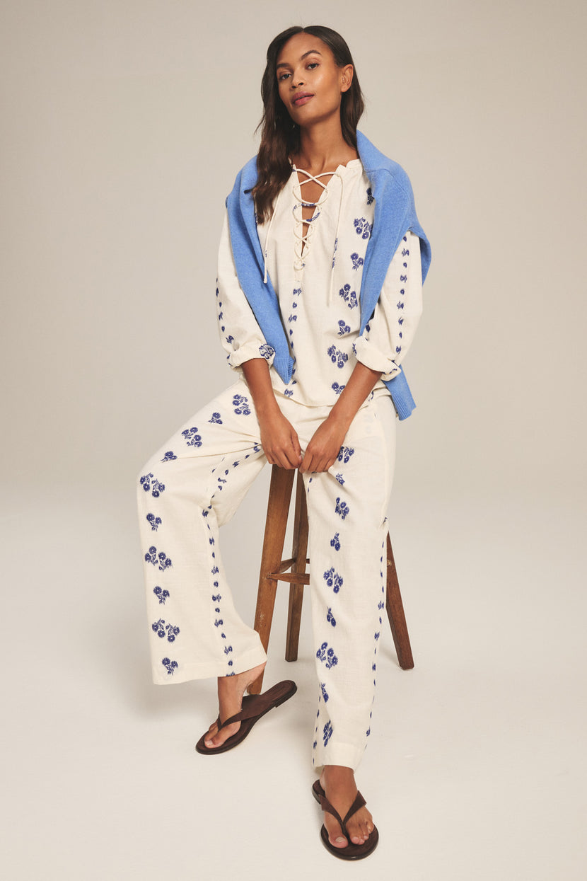 Woman wears the NELLIE PANT by Velvet by Graham & Spencer—wide-leg cotton pants with a pull-on elastic waist—paired with a cream floral top, black flip-flops, and a blue sweater draped over her shoulders while sitting on a stool.