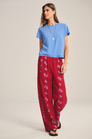 A woman stands against a plain background wearing the Velvet by Graham & Spencer NELLIE PANT, a jacquard cotton wide-leg pant with an elastic waist in red floral print, paired with a blue T-shirt, black sandals, and a necklace.