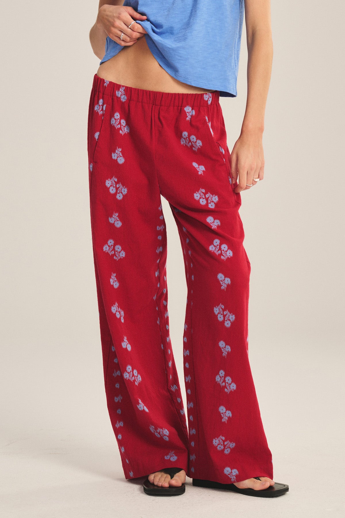 Model wearing the NELLIE PANT by Velvet by Graham & Spencer—loose red jacquard cotton wide-leg pants with blue floral prints—paired with a light blue top and black sandals, standing against a plain background.