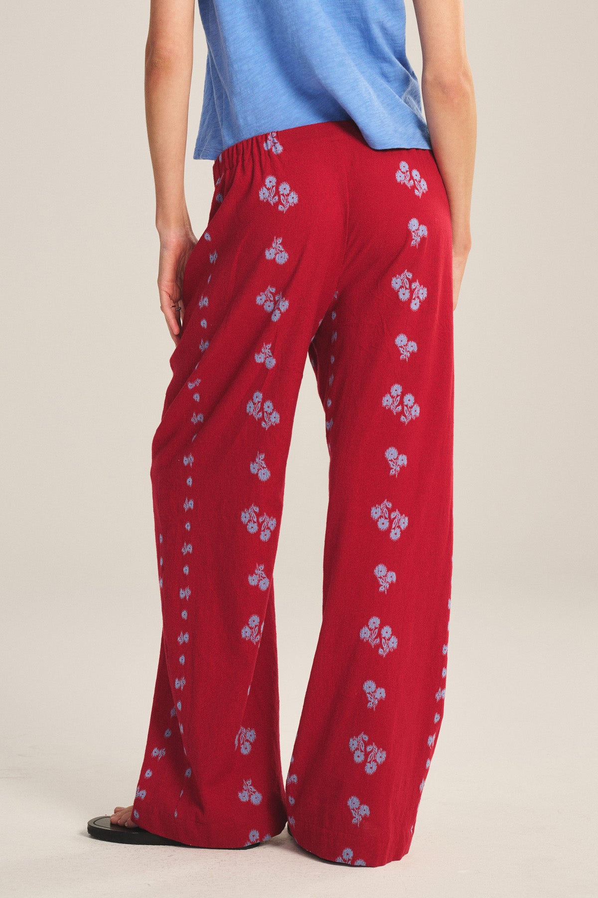 A person is seen from behind wearing the Velvet by Graham & Spencer NELLIE PANT—red, floral-patterned wide-leg pants with an elastic waist—and a blue shirt.-41089037009089