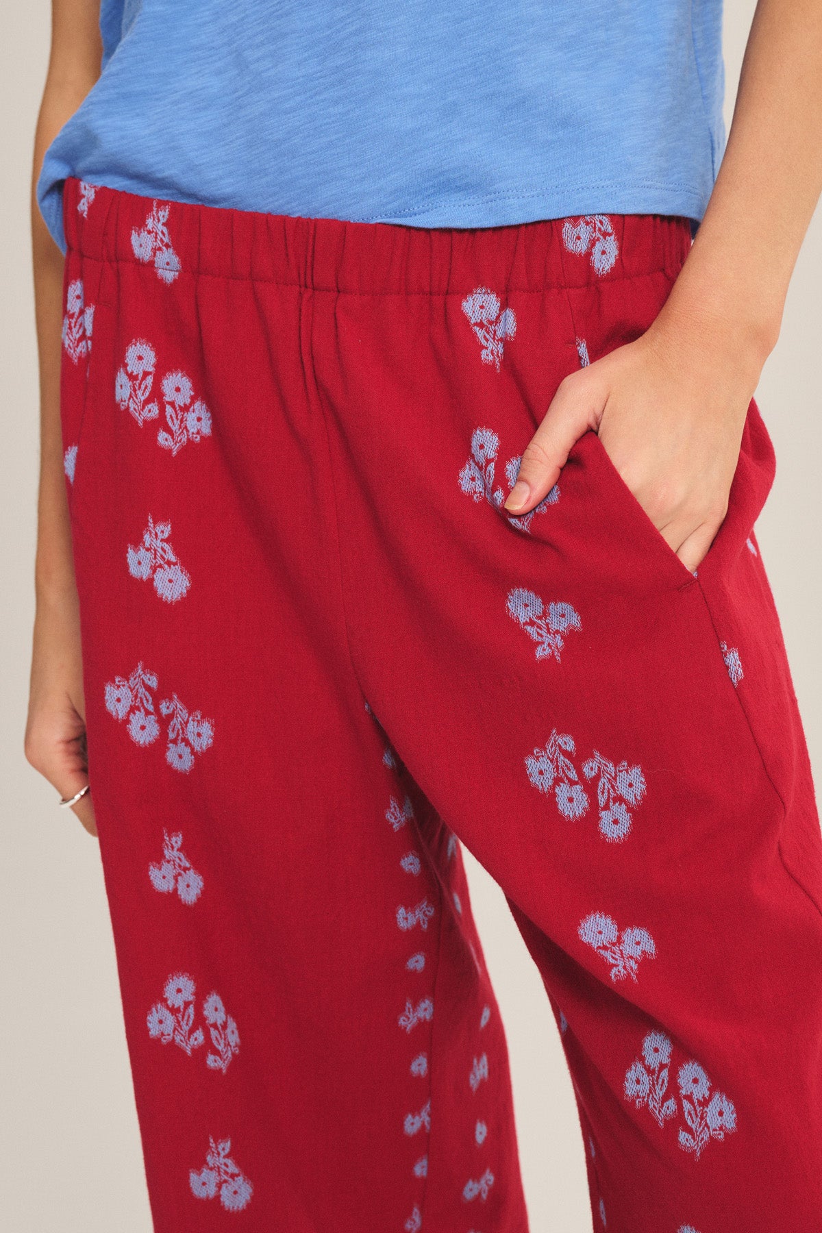 Model wearing the NELLIE PANT by Velvet by Graham & Spencer—a red wide-leg pant with a blue floral pattern and elastic waist—styled with a blue shirt, standing with one hand in their pocket.
