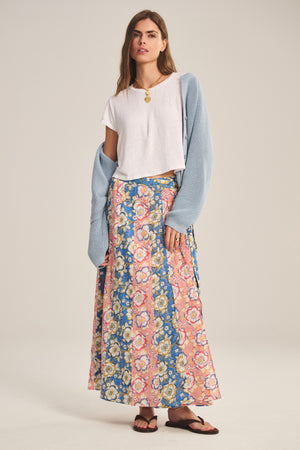 A woman wears the NICOLETTE SKIRT by Velvet by Graham & Spencer—a printed cotton maxi skirt with pink and blue patterns—paired with a white t-shirt, light blue cardigan over her arms, and black sandals.