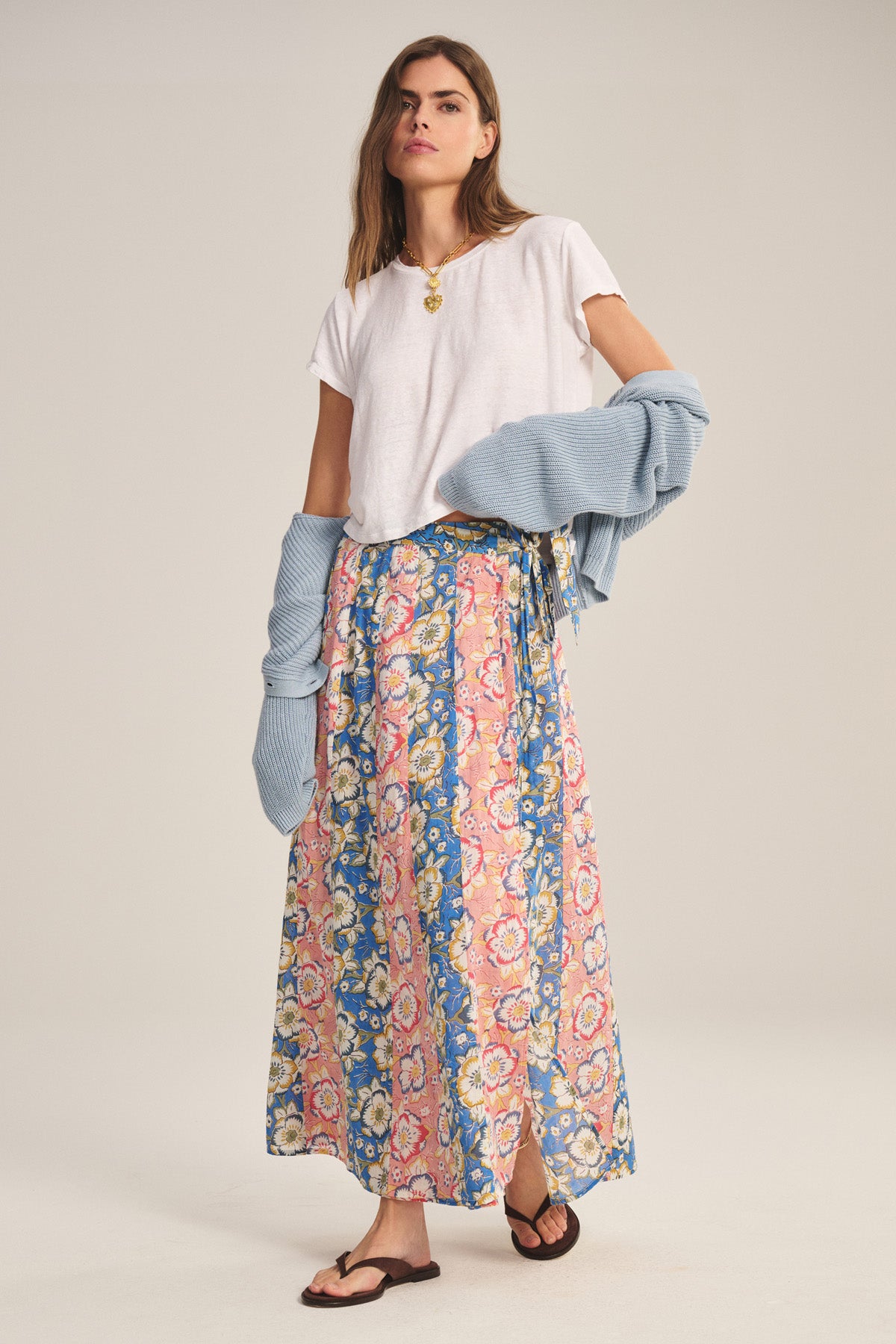A woman wears the NICOLETTE SKIRT by Velvet by Graham & Spencer—a blue, pink, and yellow printed cotton maxi skirt—paired with a white t-shirt, light blue sweater draped over her arms, and brown sandals.-41008639508673