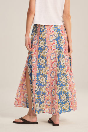 A person wearing a white sleeveless top, brown sandals, and the NICOLETTE SKIRT by Velvet by Graham & Spencer—a floral cotton maxi skirt with pink and blue patterns and a stylish side slit.