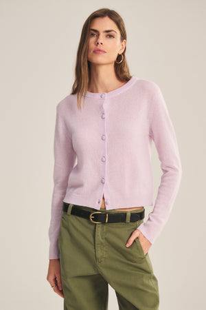 A person wears the NOLIE CASHMERE CARDIGAN by Velvet by Graham & Spencer in light purple, paired with olive green pants and a black belt, standing against a plain background.